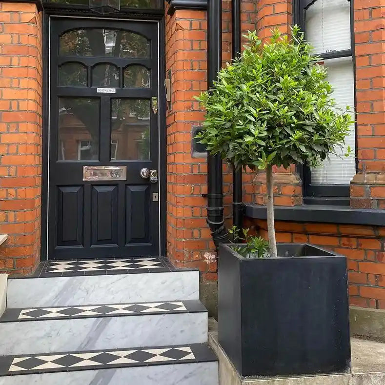 A black front door with a silver mail slot and handle on a red brick building. Patterned tiles cover the steps, and an IDEALIST Lite Contemporary Flower Box Planter sits to the right of the entrance, holding a leafy tree.
