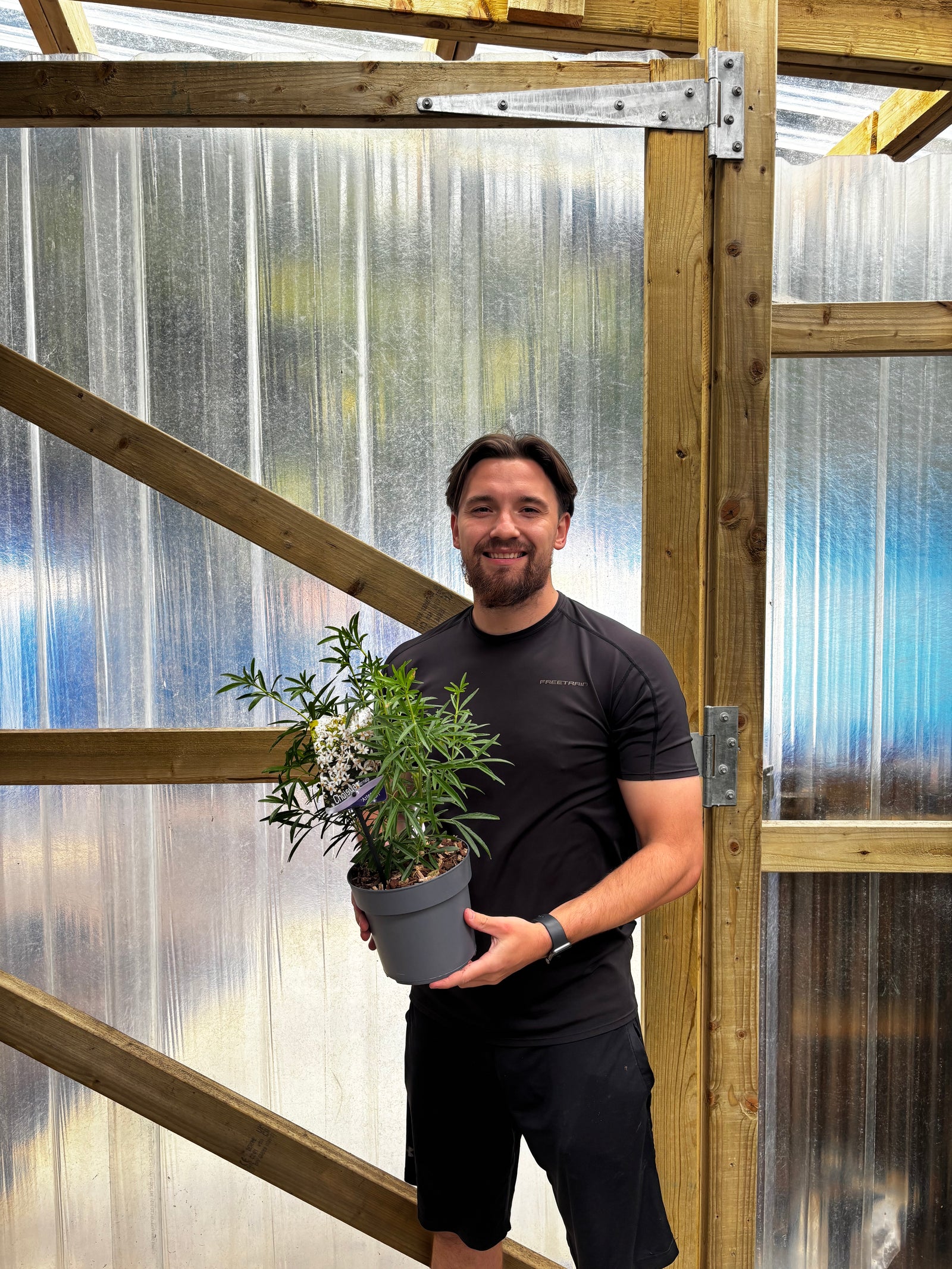 A man in a black hoodie smiles at the camera while holding a potted Choisya x dewitteana 'Aztec Pearl' 9cm / 2L, an evergreen shrub with fragrant white flowers. Shelves with more potted plants can be seen in the background.