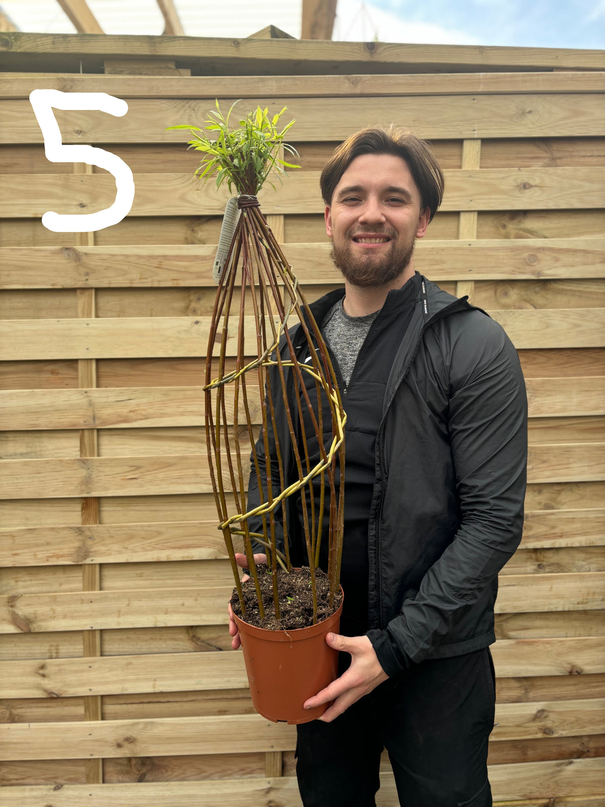 A man in a black jacket smiles while holding a Salix Living Braided Willow Tree Sculpture (80-90cm) with a tall, woven wooden frame. The number 5 appears in white at the top left, with a wooden fence in the modern garden background.