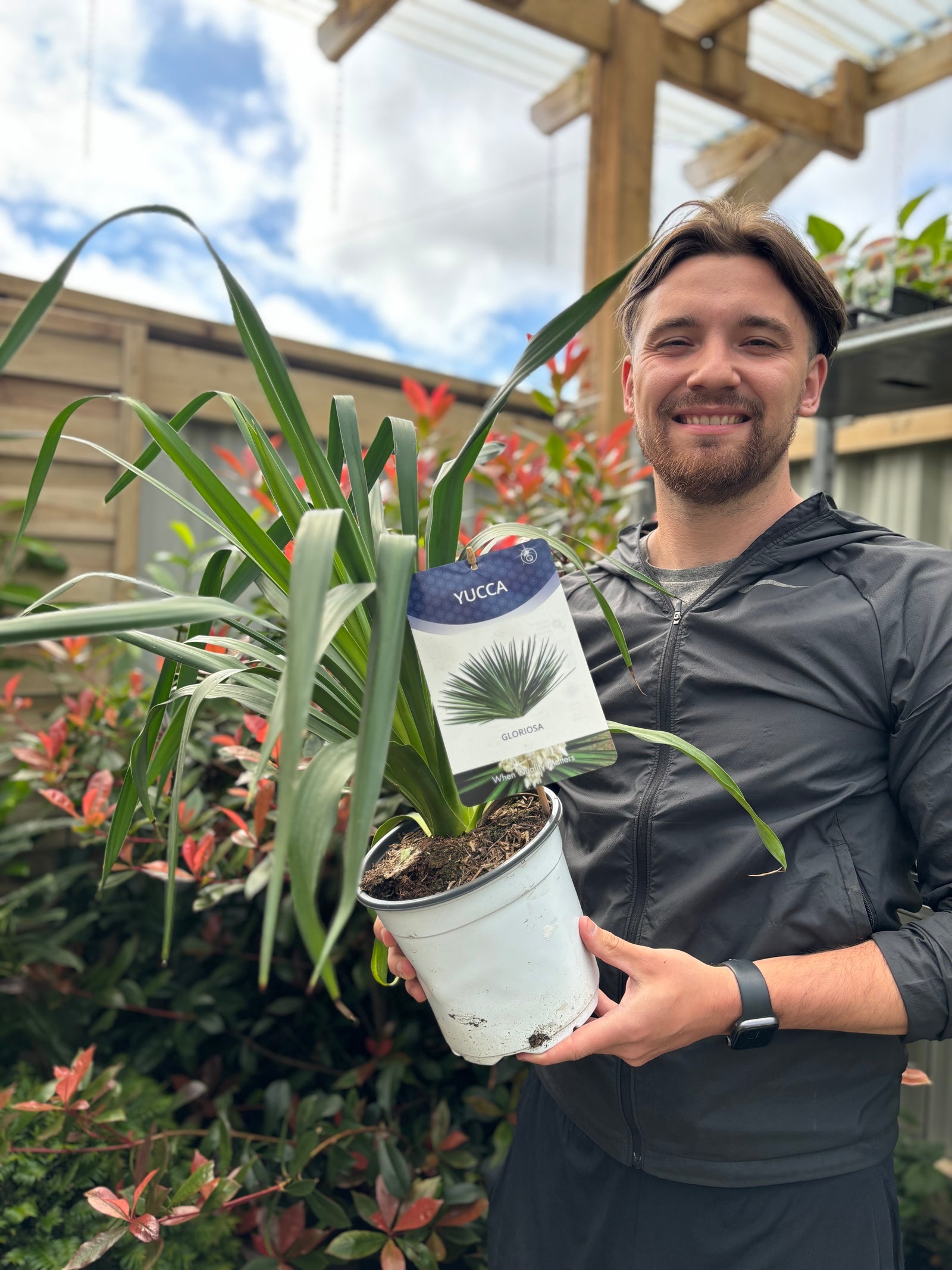 A smiling person holds a Yucca 'Gloriosa' 3L in a white pot at a garden center, surrounded by plants under a partly cloudy sky. This evergreen shrub is admired for its striking architectural form.