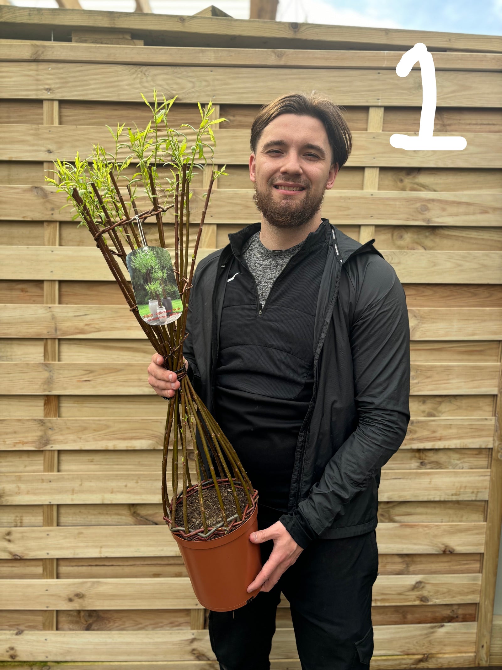 A smiling man in black sportswear holds a Salix Living Braided Willow Tree Sculpture (80-90cm) in a pot before a wooden fence, adding interest to any contemporary garden. A large white “1” appears in the top right corner.