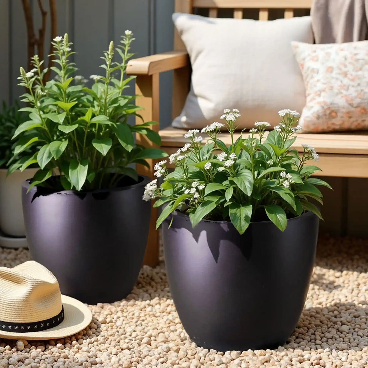 Two leafy green plants with small white flowers grow in stylish IDEALIST Lite 50cm Large Garden Planters, set on light stones. A straw hat and a wooden bench with beige and patterned cushions add charm in the background.
