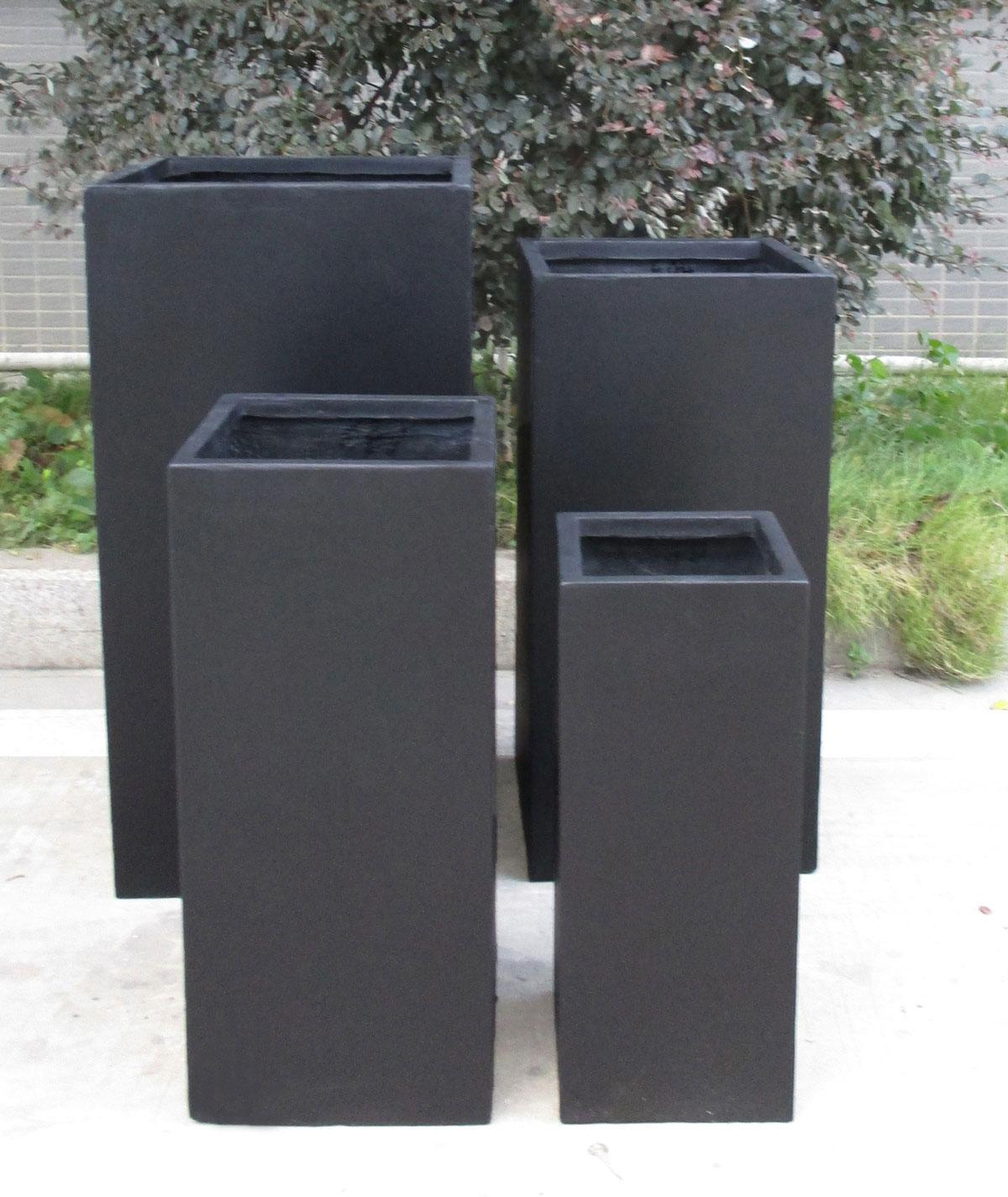 Four IDEALIST Lite Contemporary Light Concrete Tall Square Planters (H70 L33 W33 cm, 79L) are arranged outdoors on concrete, with greenery in the background. These frost-proof black pots add modern style to patios or gardens.