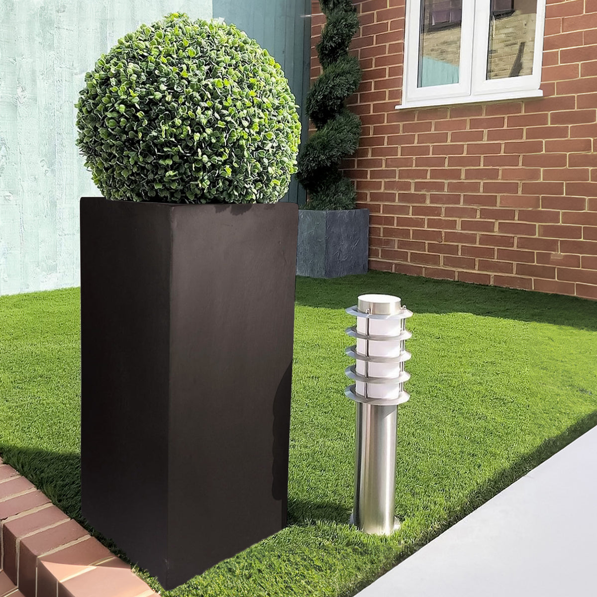 A modern garden features the IDEALIST Lite Contemporary Light Concrete Tall Square Planter (H70 L33 W33 cm, 79L) with a rounded topiary bush, beside a silver outdoor light on a manicured lawn bordered by a red brick wall and house.