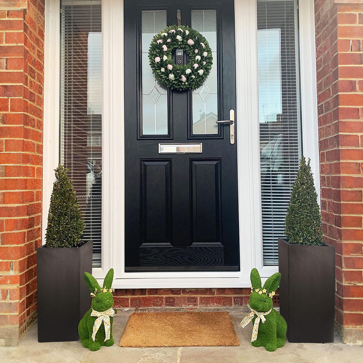 A black front door with a wreath is flanked by two IDEALIST Lite Contemporary Light Concrete Tall Square Planters. Two green bunny figures sit by a doormat, all set against a red brick wall.