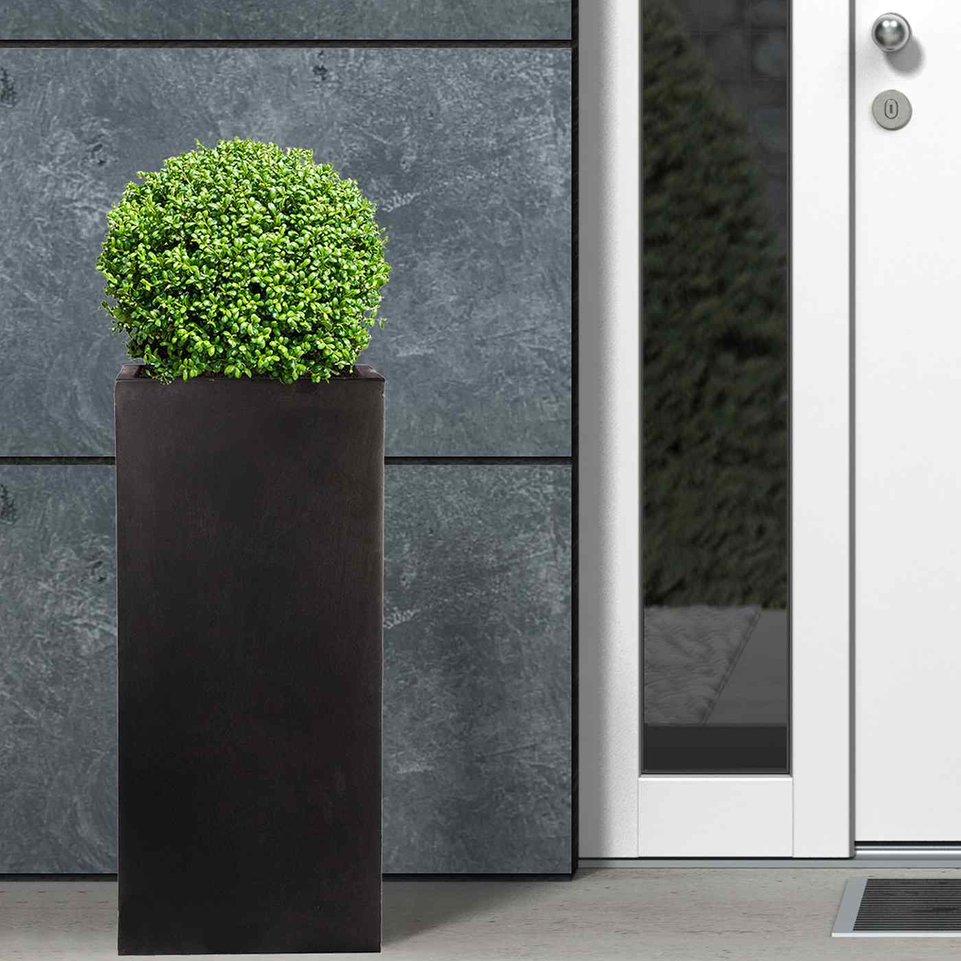 A modern black door with square windows is flanked by two IDEALIST Lite Faux Lead Tall Square Planters (50x21x21 cm, 22L) for a contemporary entrance, set in a brick wall with a small mat for a durable, stylish welcome.