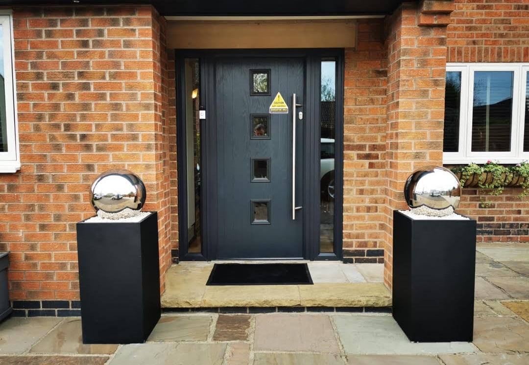 A modern black door with square windows is flanked by two IDEALIST Lite Faux Lead Tall Square Planters (50x21x21 cm, 22L) for a contemporary entrance, set in a brick wall with a small mat for a durable, stylish welcome.