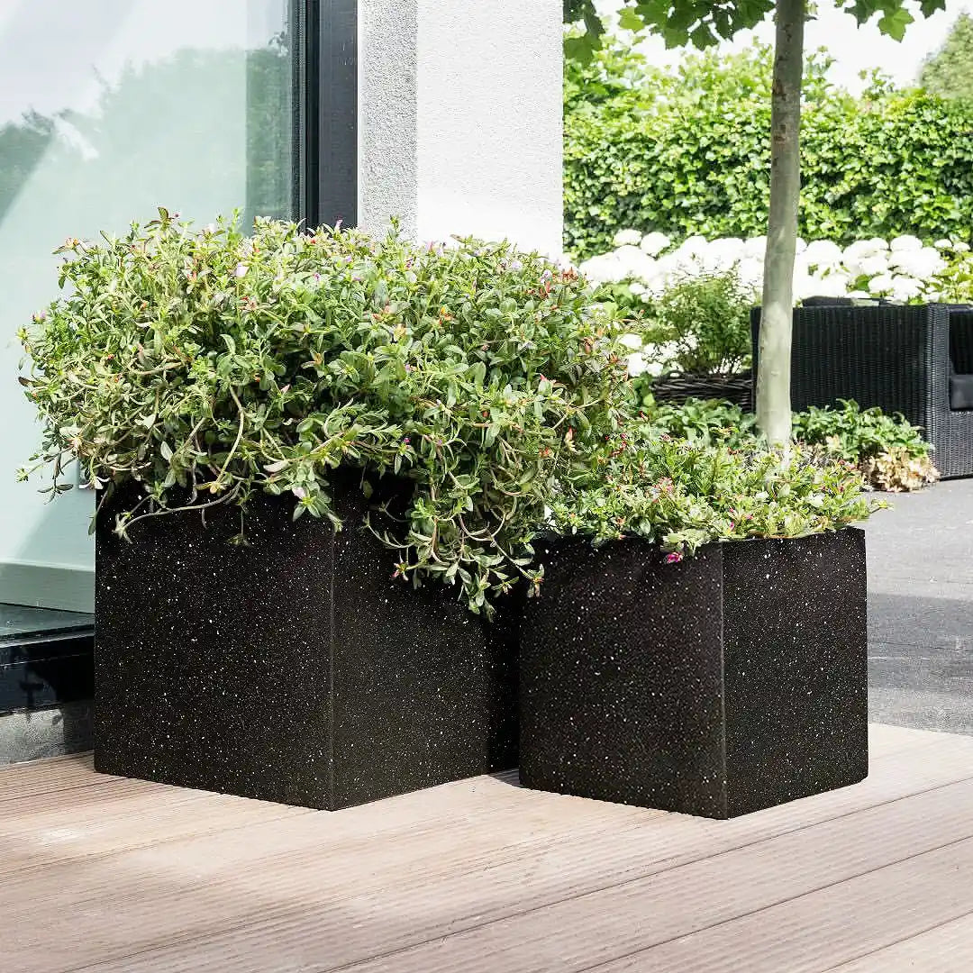 Two IDEALIST Lite Contemporary Flower Box Square Garden Planters (Light Concrete, H30 L30 W30 cm, 28L) with green leafy plants sit on a wooden deck near a glass door, with a tree and more greenery in the modern outdoor background.