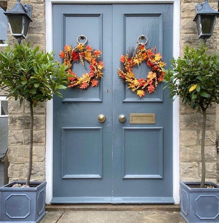 A pair of blue double doors with autumn wreaths, flanked by IDEALIST Lite Chelsea Flower Box Square Garden Planters with potted trees. Black lanterns hang on both sides of the entrance.
