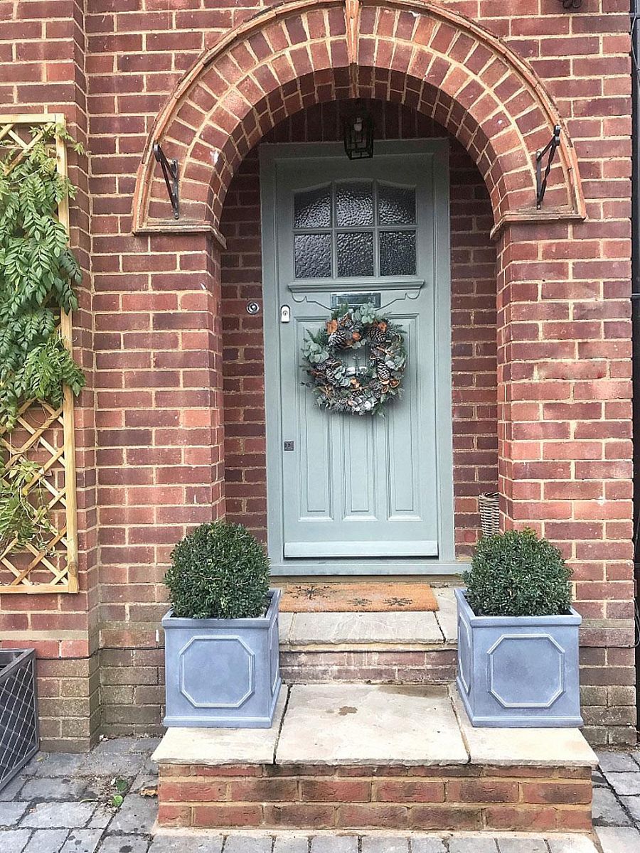 A light gray front door with a festive wreath sits under a brick archway. Two IDEALIST Lite Chelsea Flower Box Square Garden Planters with green shrubs flank the steps, and a vine climbs a trellis to the left of the doorway.