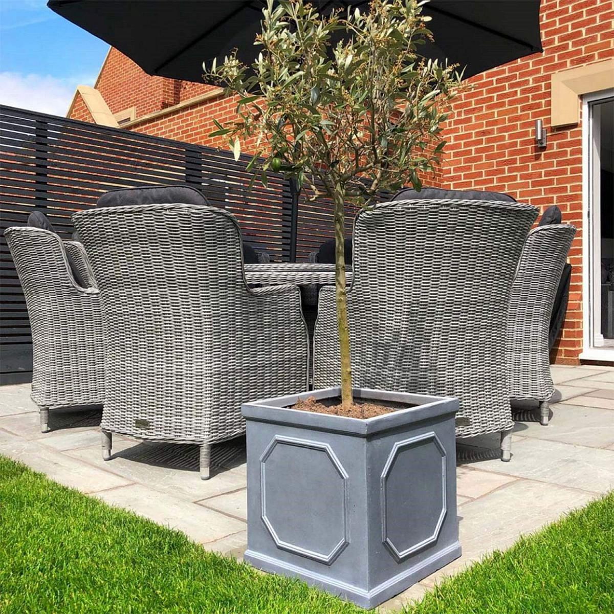 An IDEALIST Lite Chelsea Flower Box Square Garden Planter (W30 H30 L30 cm, 27L, Faux Lead Light Stone) sits on a patio with wicker chairs and table, brick wall, black fence, green grass, and an umbrella shading this cozy outdoor retreat.