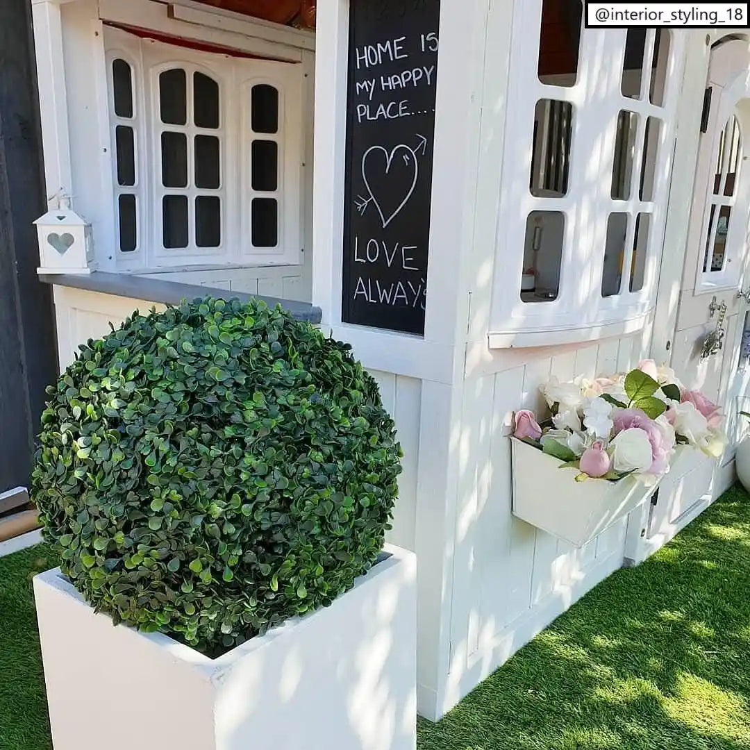 An IDEALIST Lite Contemporary Flower Box Square Garden Planter (Faux Lead Concrete, 40x40x40cm, 65L) with drainage sits on artificial grass near a white playhouse, a chalkboard reading &quot;HOME IS MY HAPPY PLACE... LOVE ALWAYS,&quot; and white-pink flowers.