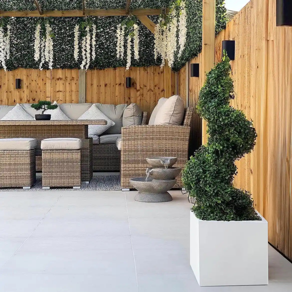 A modern outdoor seating area with wicker furniture, beige cushions, and an IDEALIST Lite Contemporary Square Garden Planter (faux lead concrete, 65L) displaying a green topiary, plus hanging white flowers and a lit wooden fence for stylish garden decor.