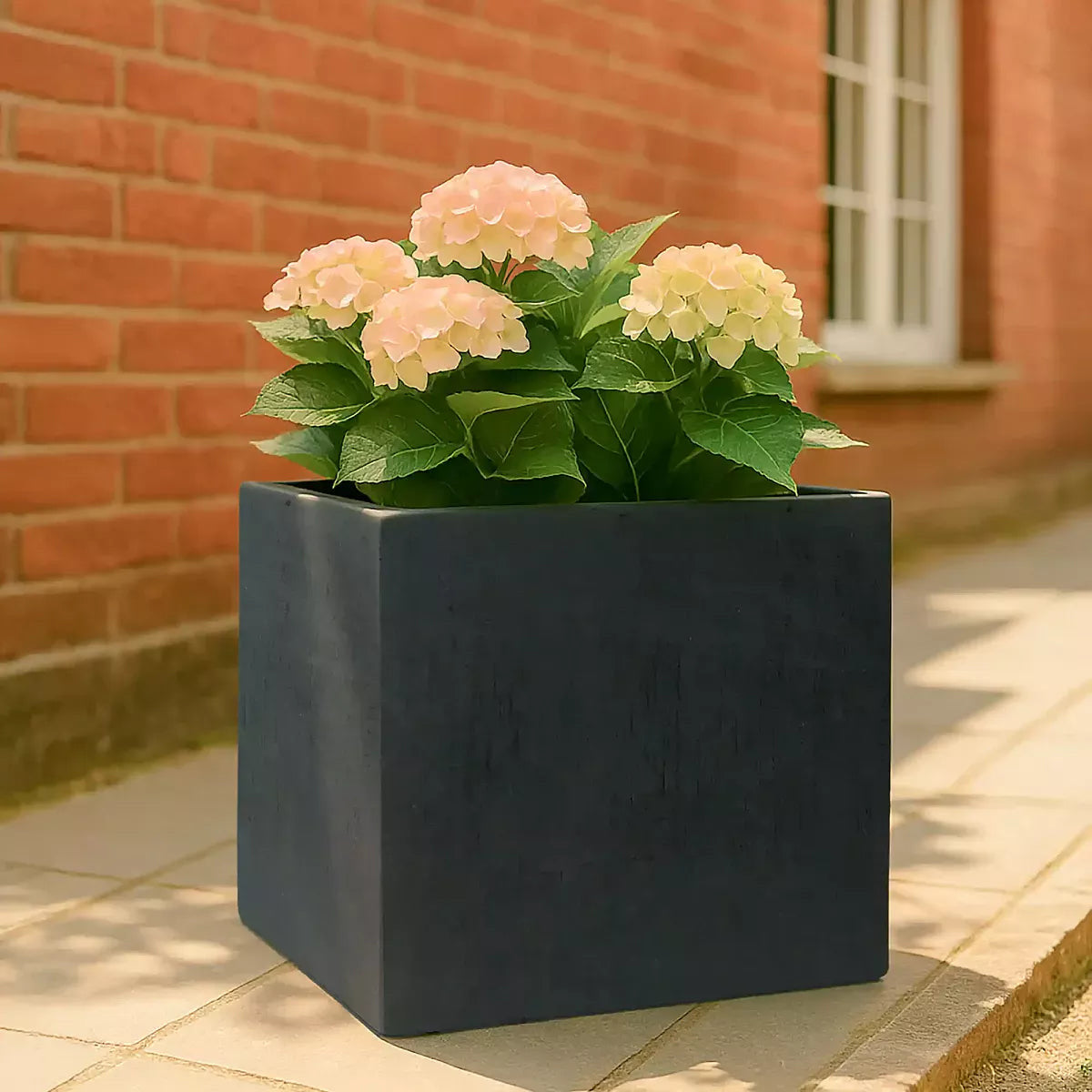 A black front door with a silver mail slot and handle on a red brick building. Patterned tiles cover the steps, and an IDEALIST Lite Contemporary Flower Box Planter sits to the right of the entrance, holding a leafy tree.