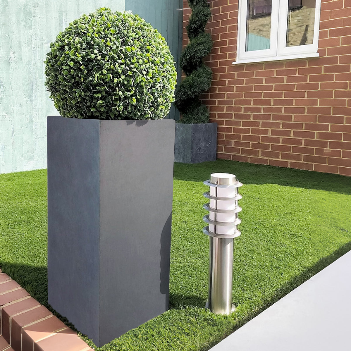 An IDEALIST Lite Contemporary Light Concrete Tall Square Planter (H70 L33 W33 cm, 79L) with a round shrub stands on a manicured lawn beside a modern outdoor stainless steel light, near a brick wall and window.