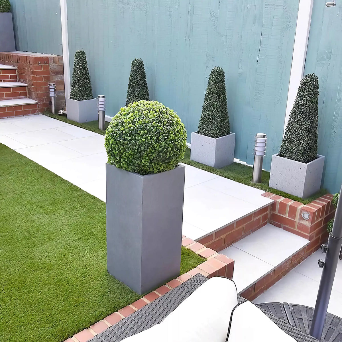 Modern backyard patio with artificial grass, white tiles, brick accents, and IDEALIST Lite Contemporary Light Concrete Tall Square Planters (H70 L33 W33 cm, 79L) holding topiary shrubs. Blue fence and silver outdoor lights highlight the space.