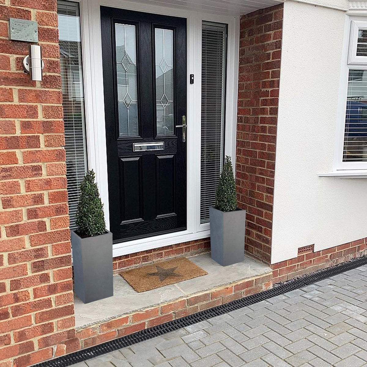 A black front door with decorative glass panels is flanked by two IDEALIST Lite Contemporary Light Concrete Tall Square Planters (H70 L33 W33 cm, 79L), trimmed bushes, a star-patterned doormat, and framed by red brick and white walls.