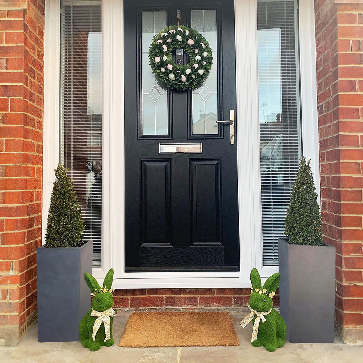 A black front door with a wreath is flanked by two IDEALIST Lite Contemporary Light Concrete Tall Square Planters. Two green bunny figures with white bows sit on either side of a doormat on the doorstep.