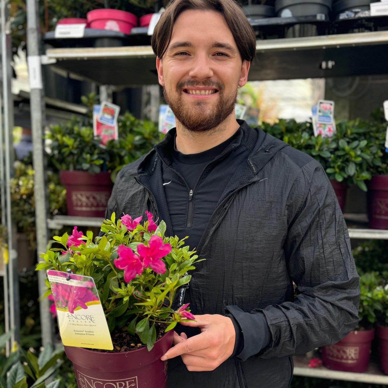 A smiling man with brown hair and a beard holds a 3L Azalea Encore 'Autumn Majesty' in a garden center, surrounded by rows of potted flowers and evergreen shrubs that promise vibrant blooms later in the season.