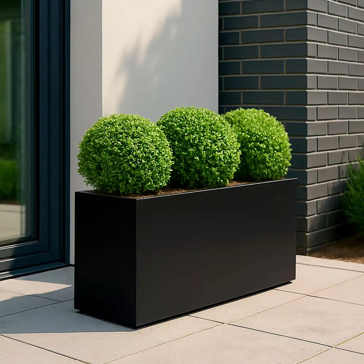 The IDEALIST Lite Contemporary Trough Garden Planter in light concrete (H30 L65 W19 cm, 37L) holds three round green bushes on a light-tiled patio beside a dark brick and white wall exterior.