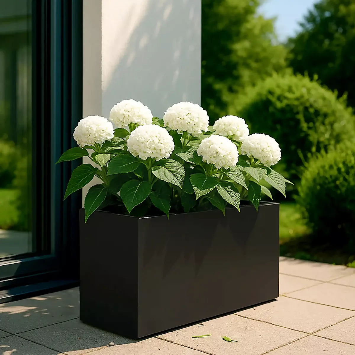 The IDEALIST Lite Contemporary Trough Garden Planter (H30 L65 W19 cm, 37L, light concrete) sits on a tiled patio near a glass door, holding blooming white hydrangeas with green leaves, with bushes and trees in the background.