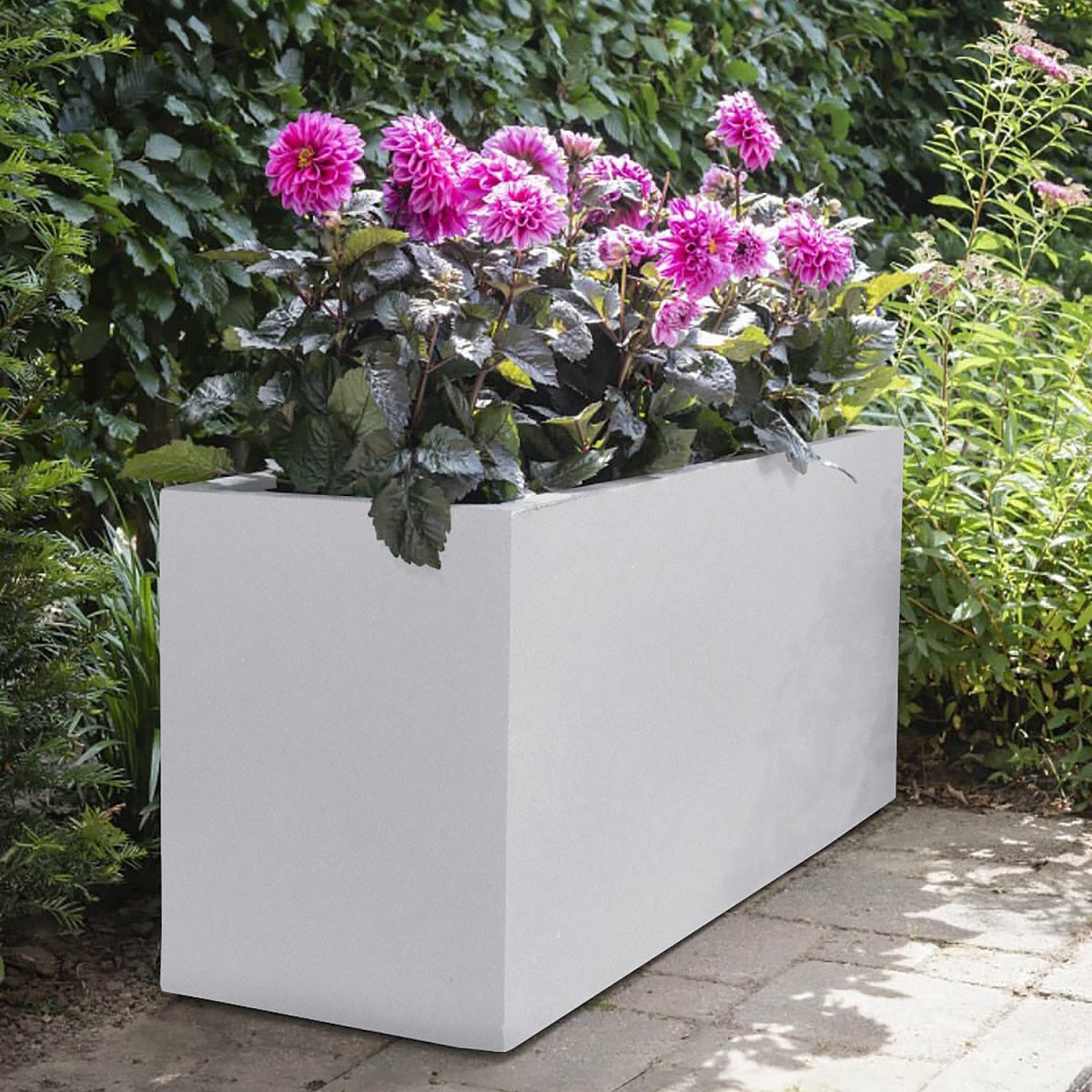 The IDEALIST Lite Contemporary Trough Garden Planter (H51.5 L100 W36 cm, 185L, light concrete) filled with blooming pink dahlias sits on a stone patio, surrounded by greenery, adding elegant garden decor to the scene.