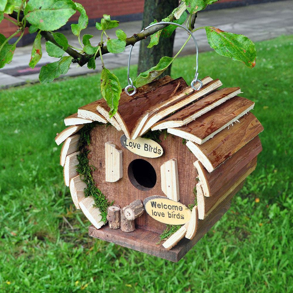 The Wooden Crafted Bird Hotel is a charming log cabin-shaped nesting box with a circular entry hole, two front signs—"Love Birds" and "Welcome love birds"—and a sturdy metal hanging wire for easy display.