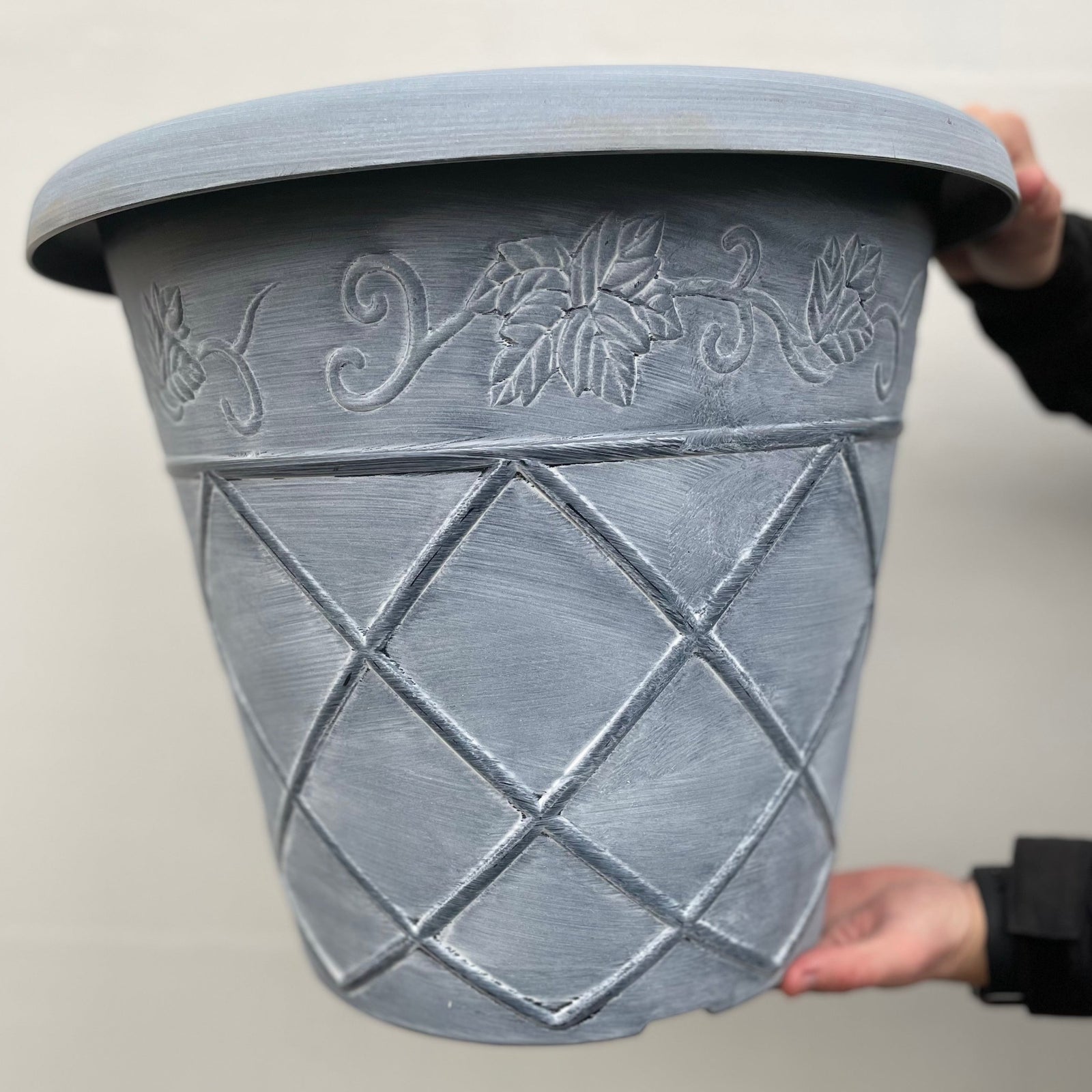 A person holds the Shabby Chic Washed Planter in light blue, a large round plastic pot with a diamond pattern on the lower half and vine and leaf designs on the top edge.