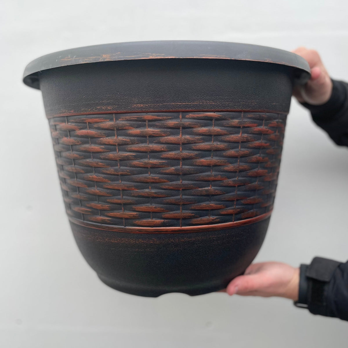 A person holds the Wicker Barrel Planter - Bronze, a large, durable plastic planter with a woven basket-like texture and reddish bronze accents, set against a plain light background.