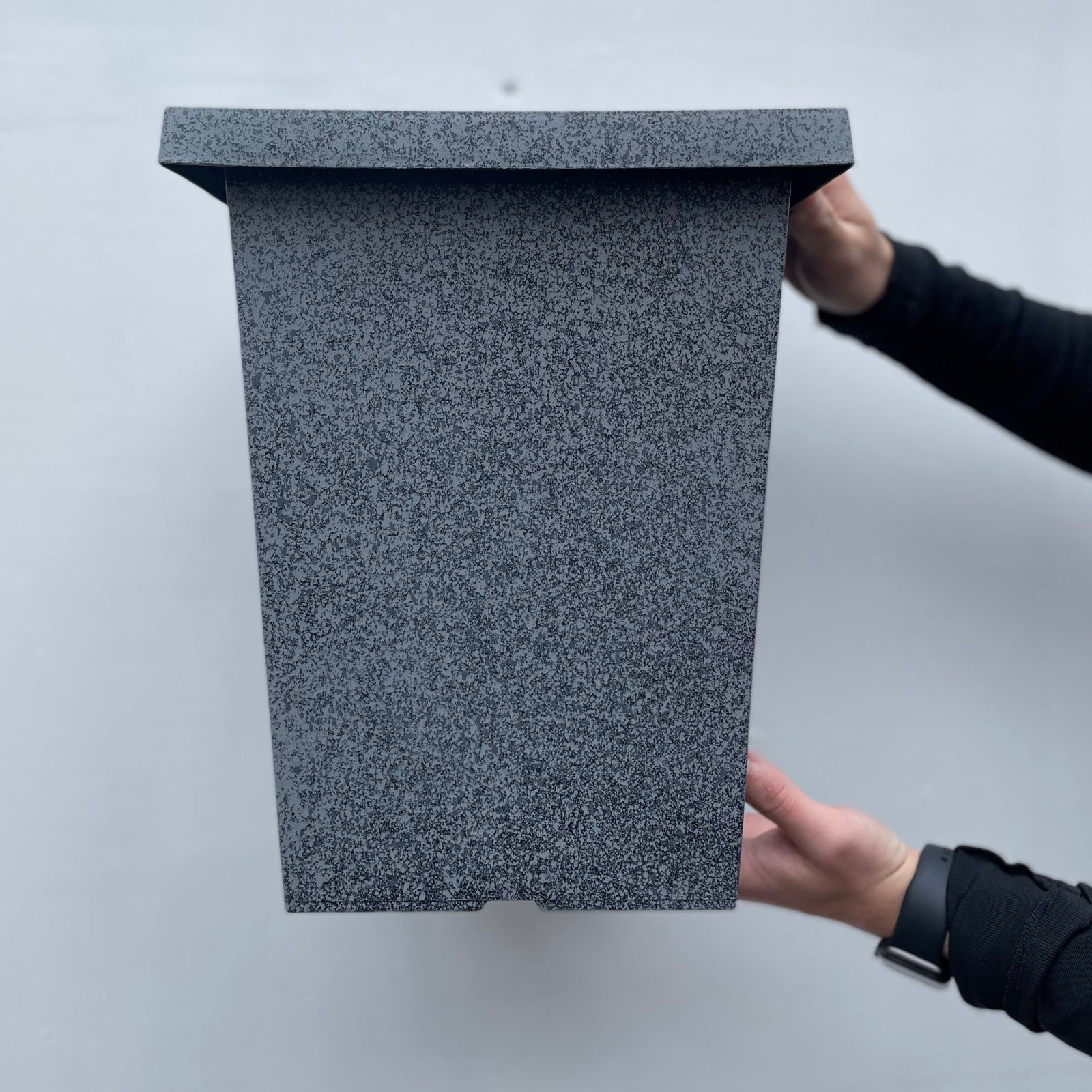 A person in black long sleeves holds the Granite Effect Finish Planter - Grey, a large rectangular box with a speckled granite finish and slanted lid, against a light gray background.