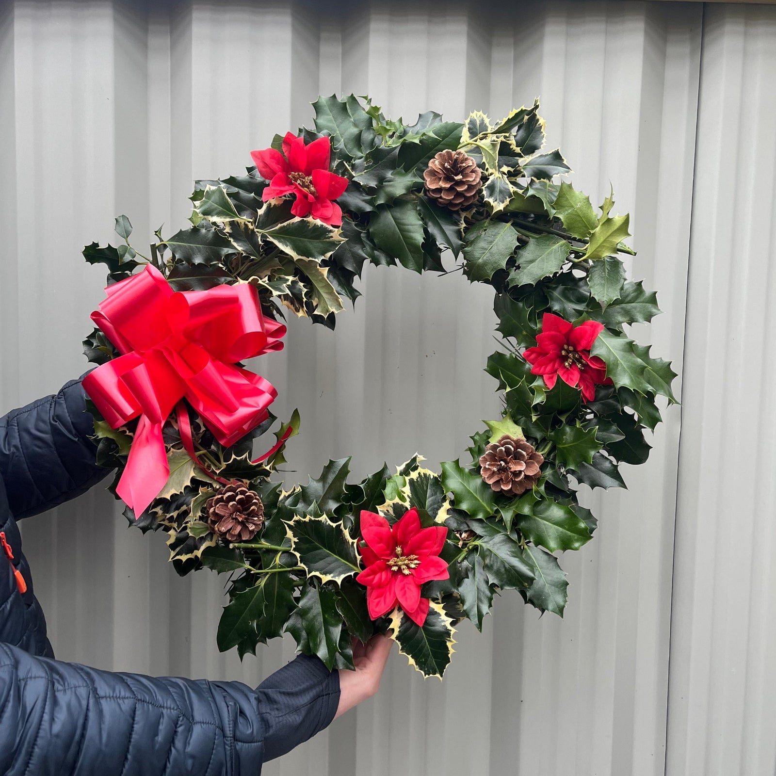 A young man in glasses and a black puffer jacket holds the Real Holly Wreath | Hand Made Wreath (45cm), a festive door decoration adorned with red bows, flowers, pinecones, and green foliage, standing in front of a corrugated metal wall.