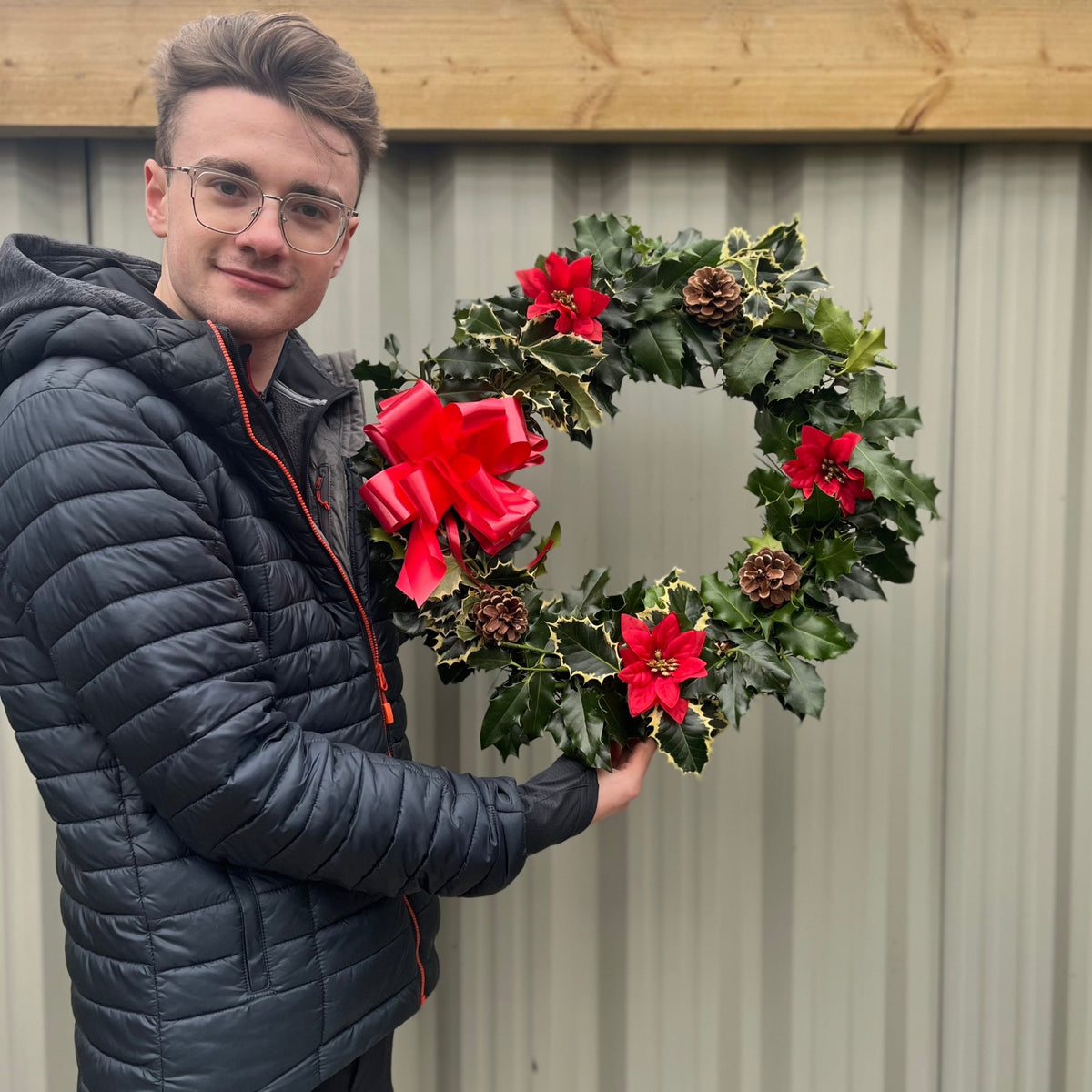 A young man in glasses and a black puffer jacket holds the Real Holly Wreath | Hand Made Wreath (45cm), a festive door decoration adorned with red bows, flowers, pinecones, and green foliage, standing in front of a corrugated metal wall.