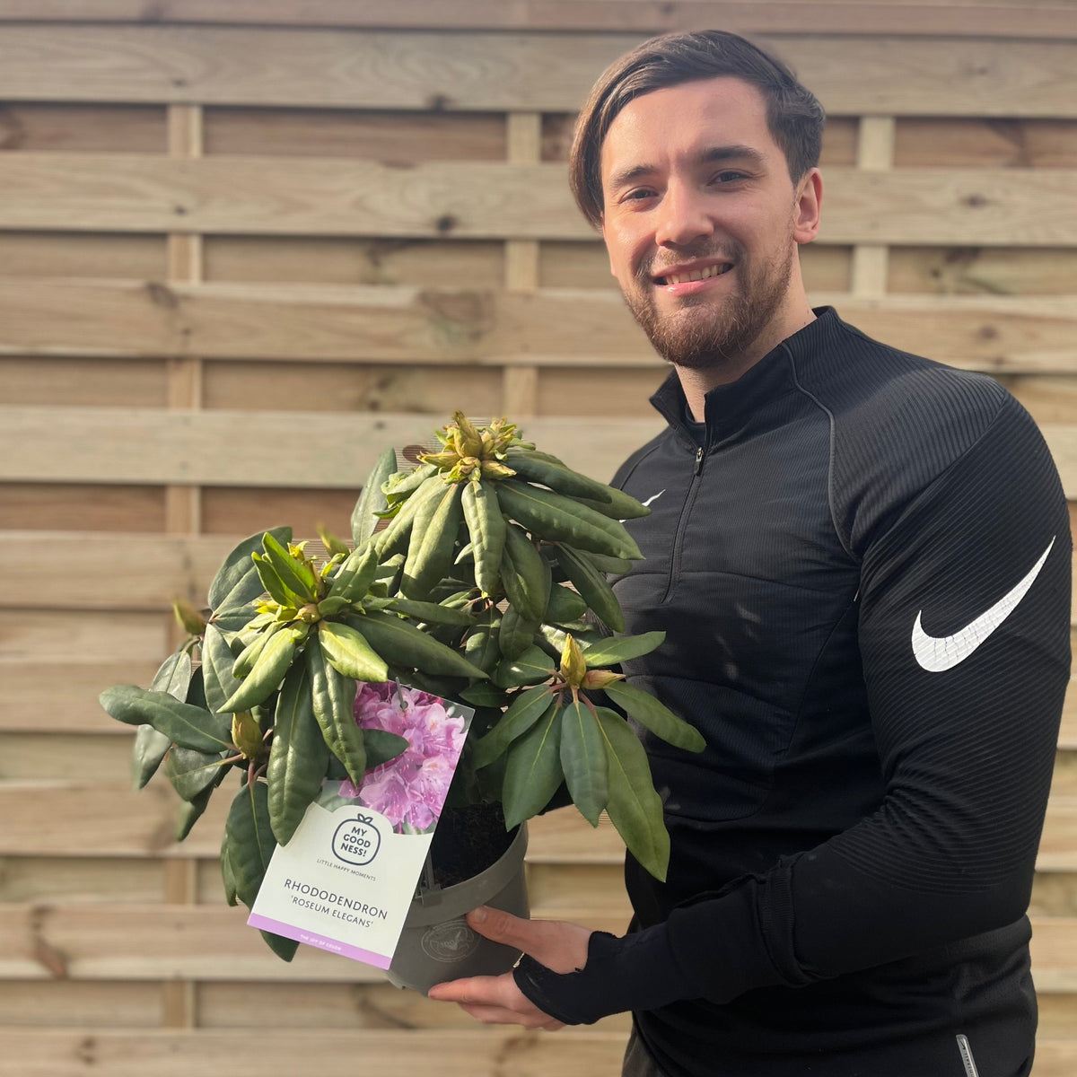 A smiling person in a black zip-up Nike top holds a Rhododendron Delta 2L / 5L with an attached label, standing in front of a wooden fence.