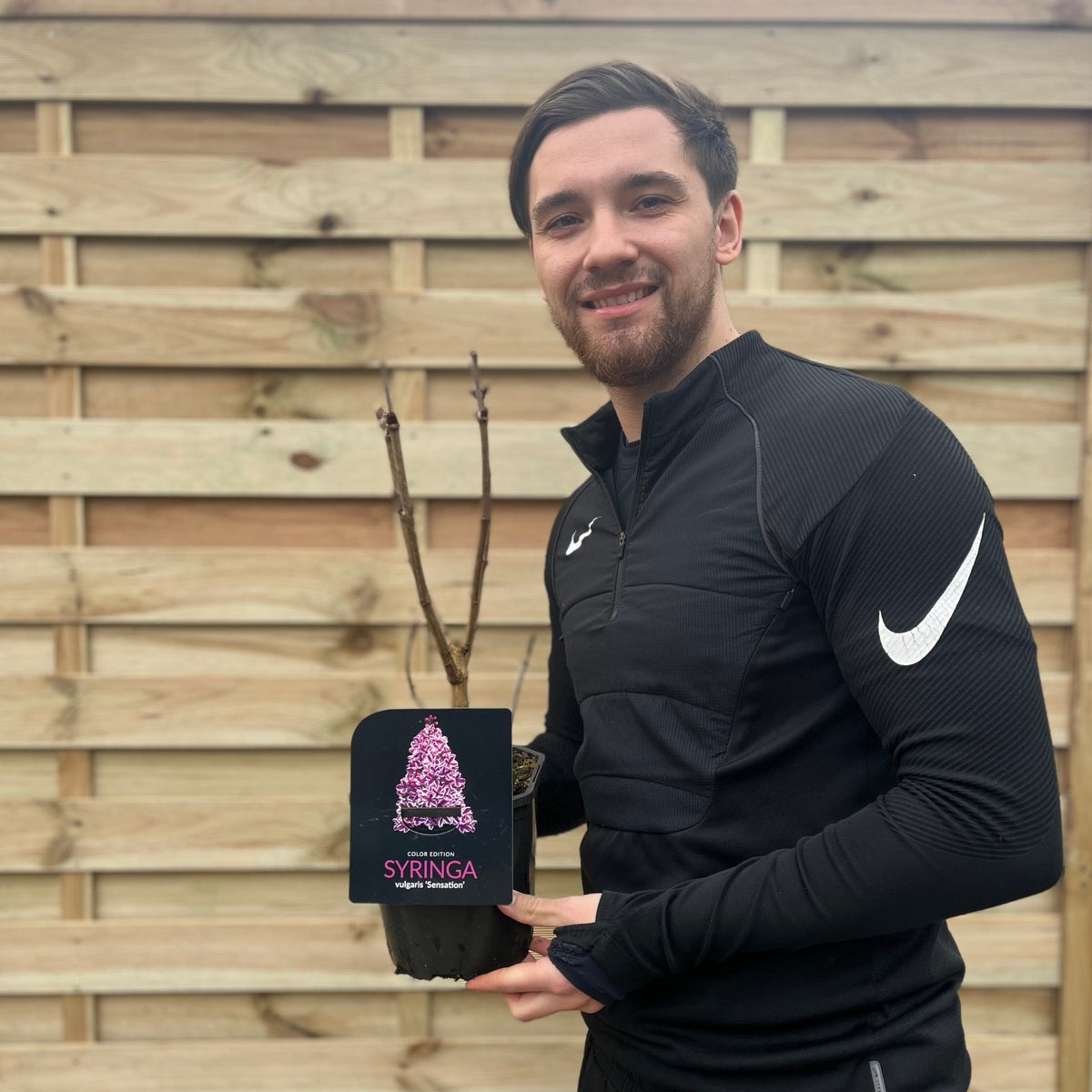 A man in a black sports outfit smiles while holding a Syringa vulgaris &#39;Sensation&#39; (Lilac) 2L in front of a wooden fence—ready to add its fragrant blooms to his garden.