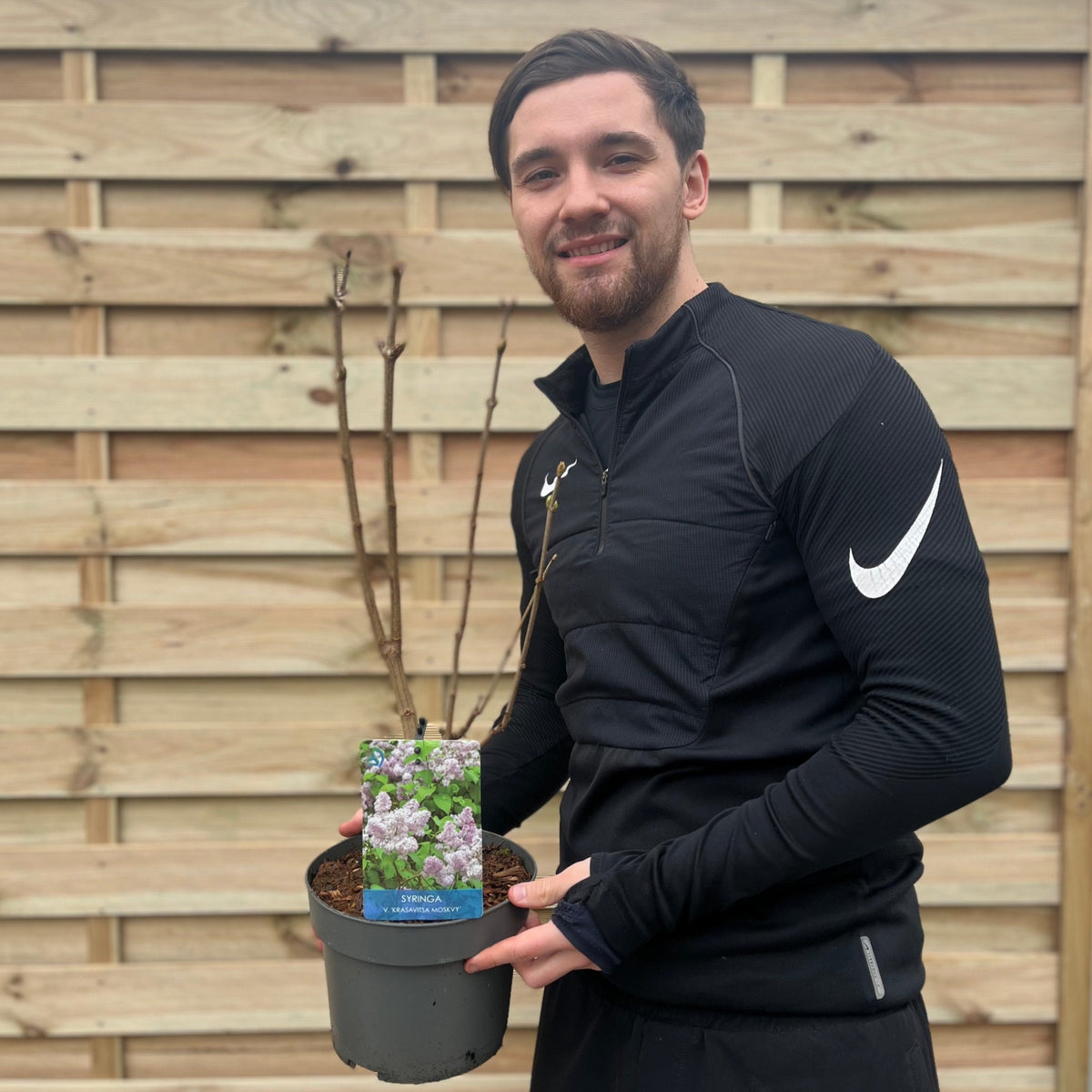 A man in black athletic wear stands before a wooden fence, smiling and holding a Syringa vulgaris &#39;Beauty of Moscow&#39; (Lilac) 3L, its label displaying fragrant white blossoms.