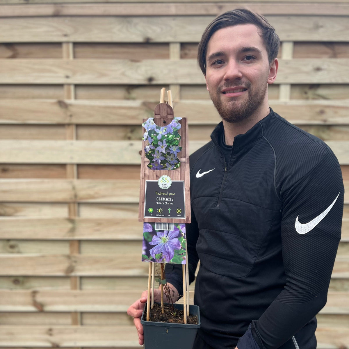 A man in a black Nike tracksuit smiles in front of a wooden fence, holding a potted Clematis &#39;Prince Charles&#39; 75cm, a compact climber with mauve-blue flowers and a label attached.