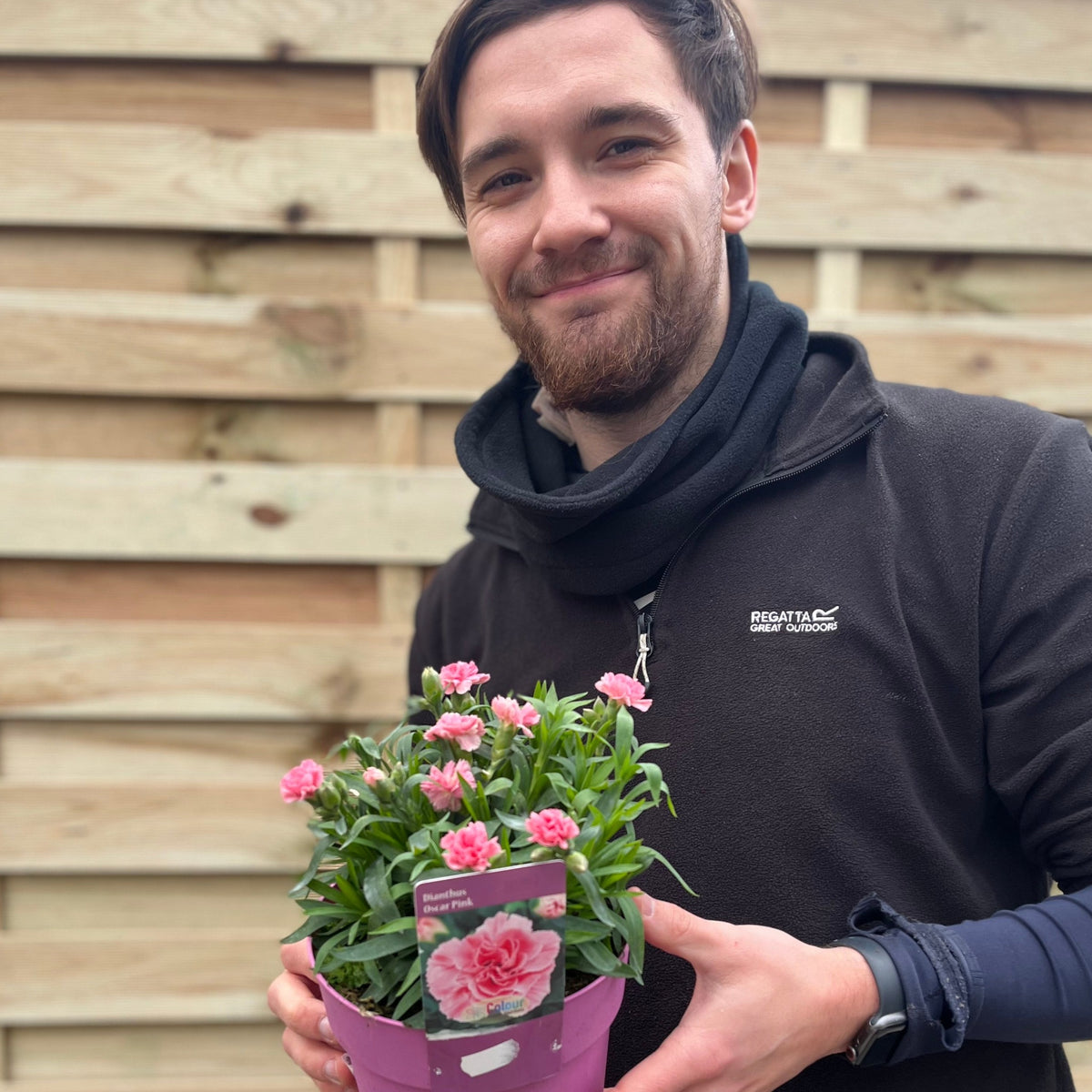 A smiling bearded person with short brown hair holds a pink pot of Dianthus &#39;Oscar Pink&#39;, a compact, drought-tolerant perennial. They stand by a wooden fence, dressed in a black jacket and scarf.