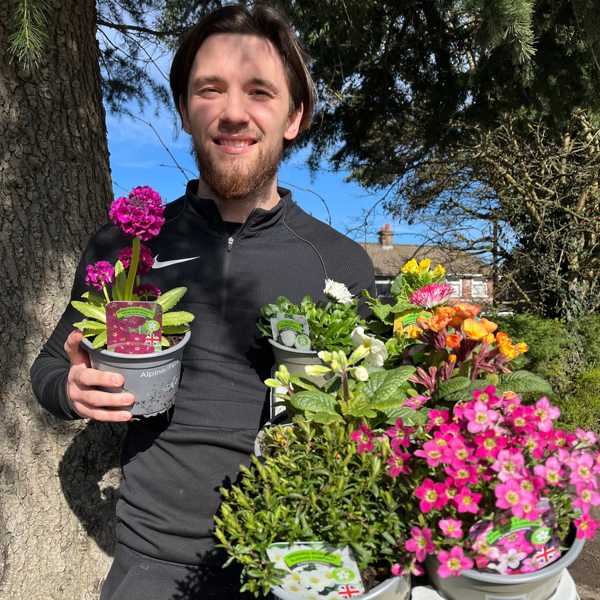 A smiling man in black stands outside on a sunny day, holding a Special Offer - Mix of 5 Spring Flowering Perennials, with garden displays, trees, and a house in the background.
