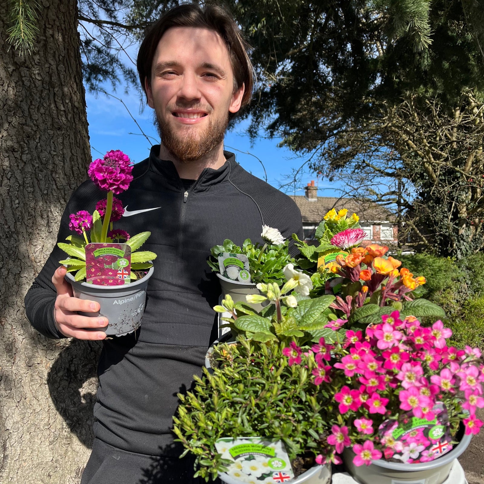 A smiling man in black stands outside on a sunny day, holding a Special Offer - Mix of 5 Spring Flowering Perennials, with garden displays, trees, and a house in the background.