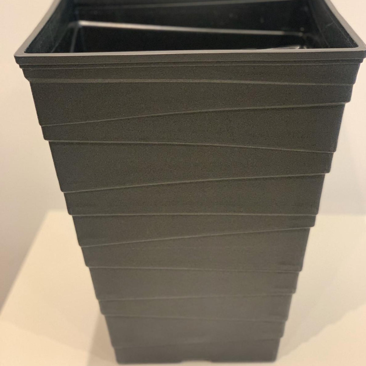 The Luxury Ripple Plastic Plant Pot - BLACK is a tall, square pot with horizontal ridges and drainage holes, shown on a light-colored surface against a plain background.