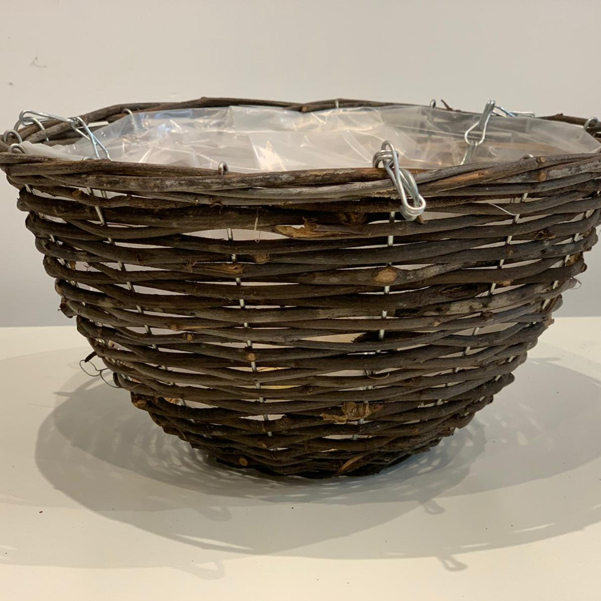 The Wicker Hanging Basket, a round woven wicker piece 28cm wide with clear plastic lining and metal clips, sits on a plain white surface against a neutral background.