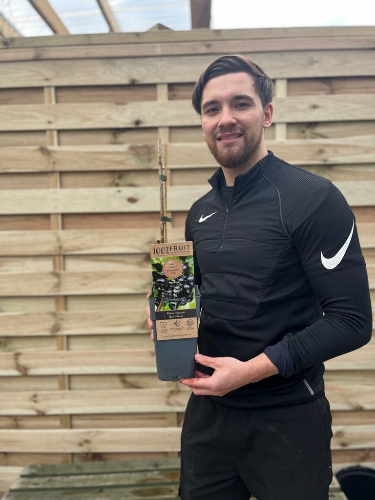A man in a black Nike tracksuit smiles outdoors, holding a 2L Blackcurrant &#39;ribes nigrum&#39; Ben Nevis bush labeled “100% FRUIT” in front of a wooden fence.