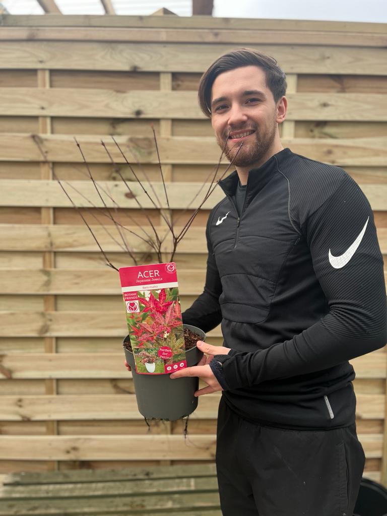 A man in a black Nike tracksuit smiles while holding an Acer palmatum Beni-maiko 3L with red leaves, standing in front of a wooden fence.
