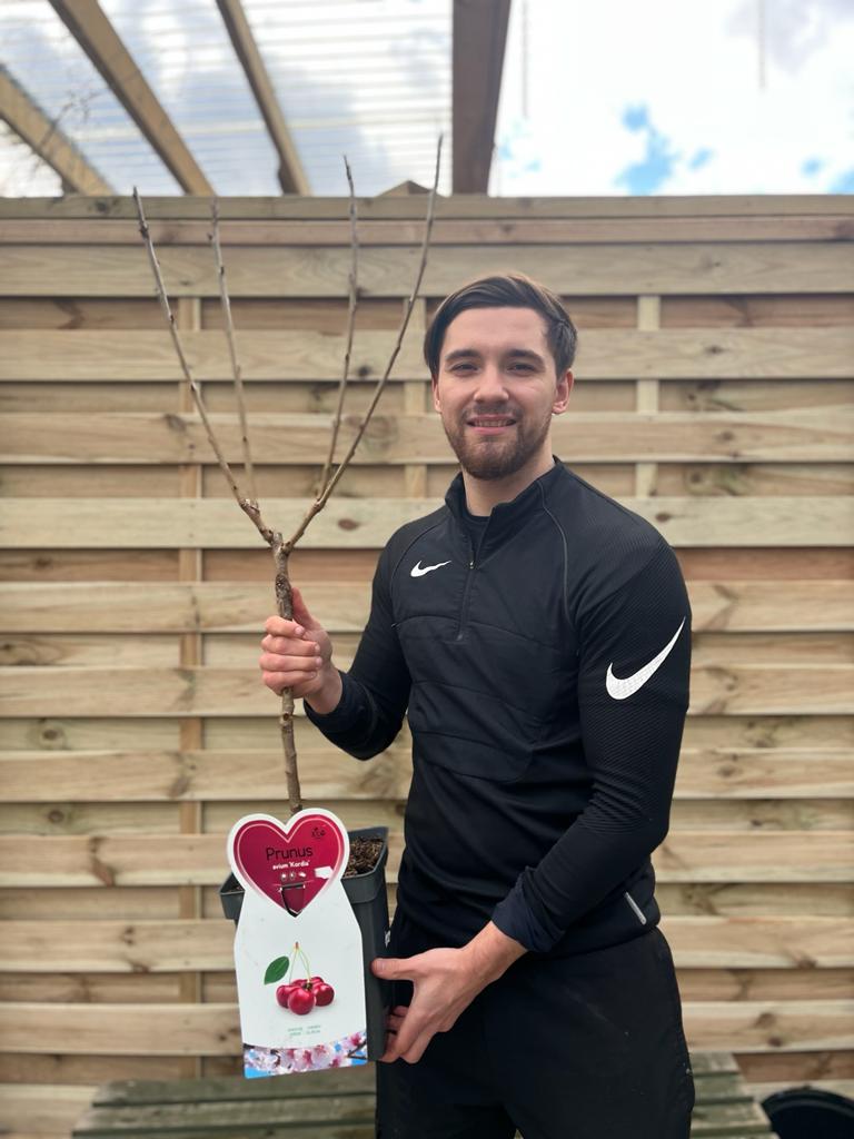 A smiling man in a black Nike tracksuit holds a Dwarf Patio Cherry Tree &#39;Kordia&#39; 1M in front of a wooden fence on a cloudy day. The pot features a label with cherries and a heart-shaped tag.