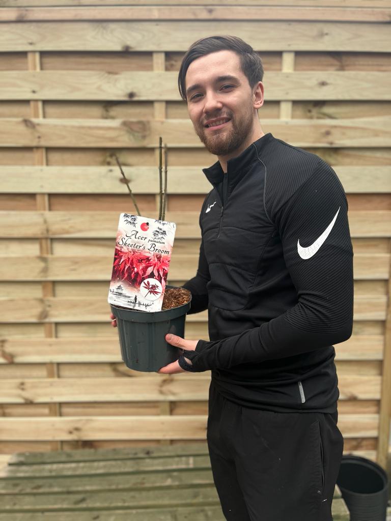 A smiling man in black athletic wear holds an Acer palmatum Skeeter&#39;s Broom 3L, a deciduous shrub with vivid red-purple leaves, in front of a wooden fence.